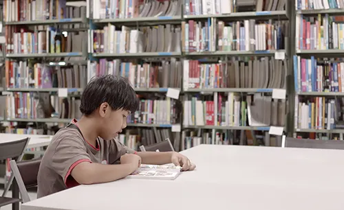A young boy reading a book at a table between tall library shelves