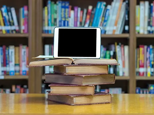 A laptop resting on a stack of books in front of library shelves