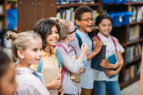A group of smiling schoolchildren with backpacks standing in front of library shelves
